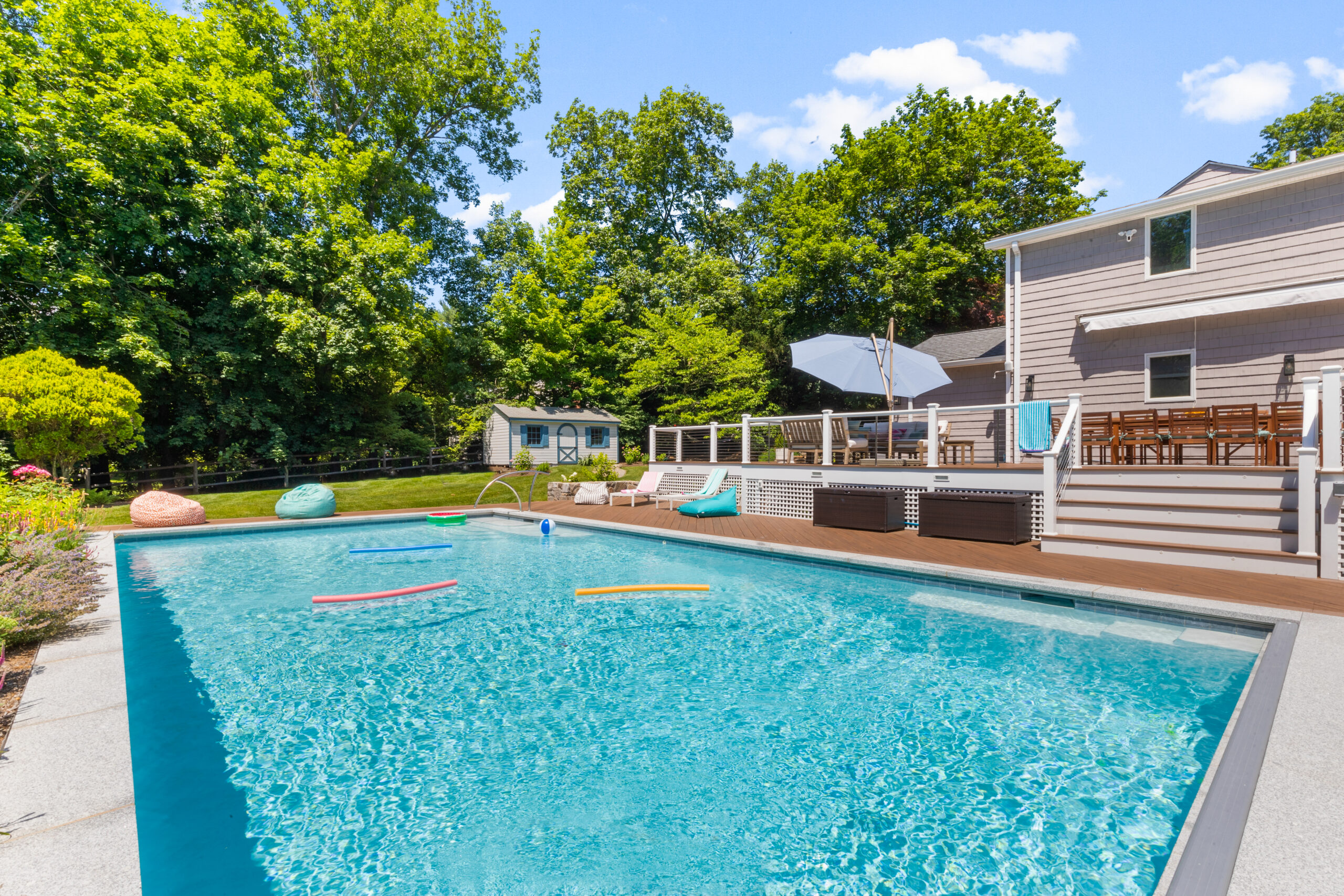 Swimming pool in the backyard along with lounge chairs, wooden deck, and seating area surrounded by lush trees.