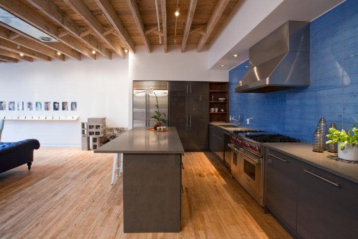 open kitchen with dark gray kitchen and stainless steel appliances and large hood and bright blue backsplash and hardwood floors and ceiling beams after renovation