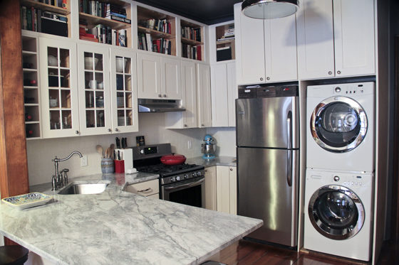 white kitchen cabinets with gray marble countertop and book shelves above see through overhead cabinets and stainless steel appliances and stackable laundry machines next to refrigerator after renovation