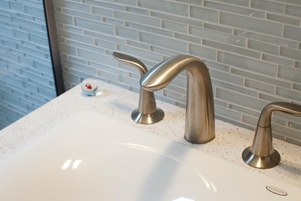 bathroom sink with brushed nickel faucets and light gray glass wall tiles after renovation