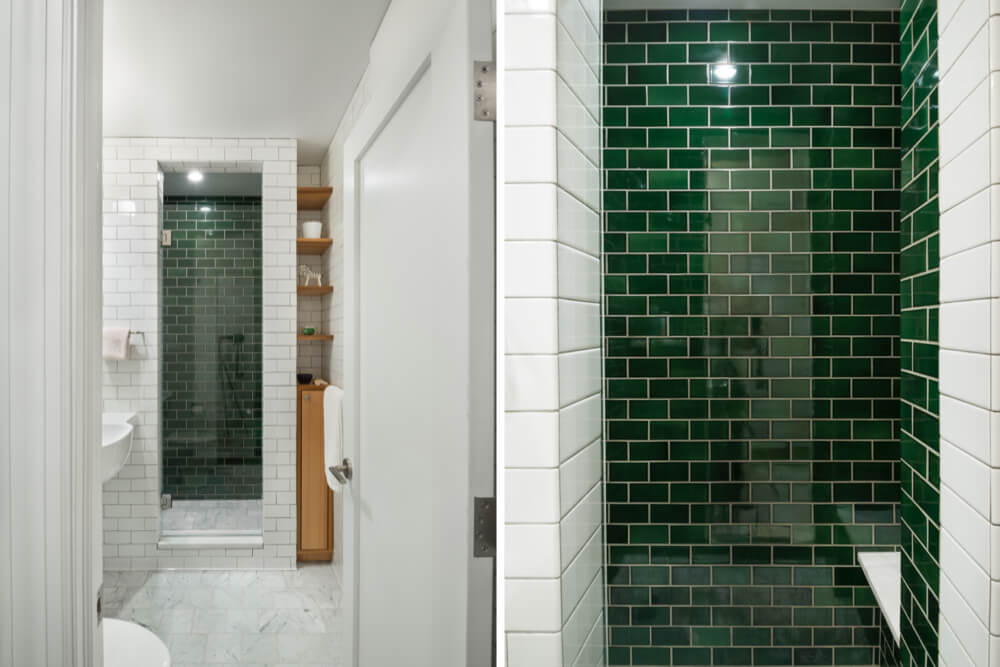 two images of white bathroom with built-in oak wood storage and white subway wall tiles and walk-in shower with dark green subway tiles on wall after renovation 