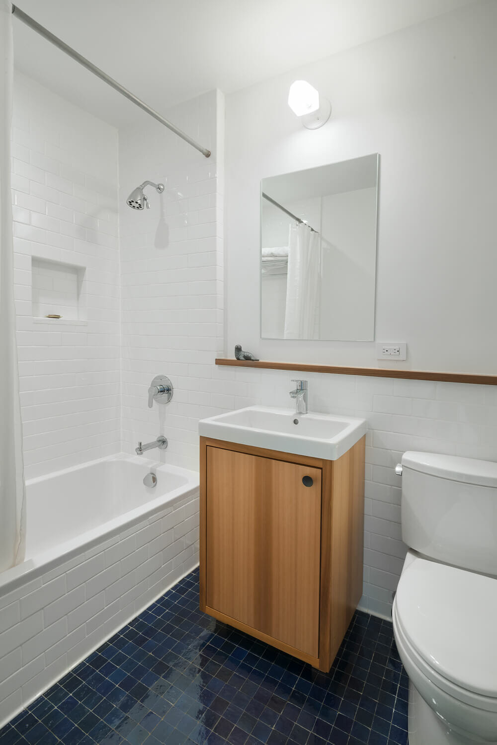 bathtub with recessed shower shelf and oak vanity with mirror and toilet and white wall tiles with wood trim and black floor tiles after renovation