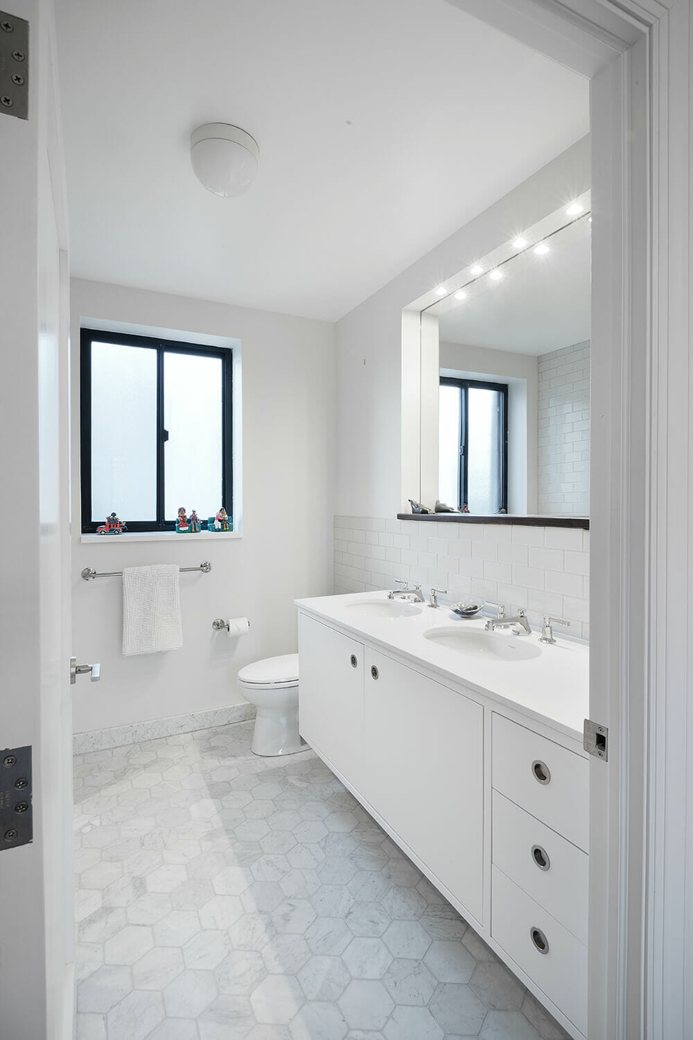 white double vanity sink with large mirror and white hexagon floor tiles and window with black frames next to toilet after renovation