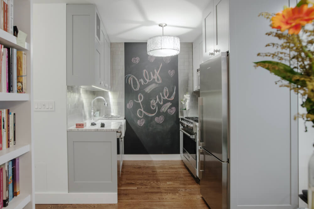 small kitchen with gray cabinets and stainless steel appliances and large chalkboard on wall and pendant light and built-in book shelves in the hallway after renovation
