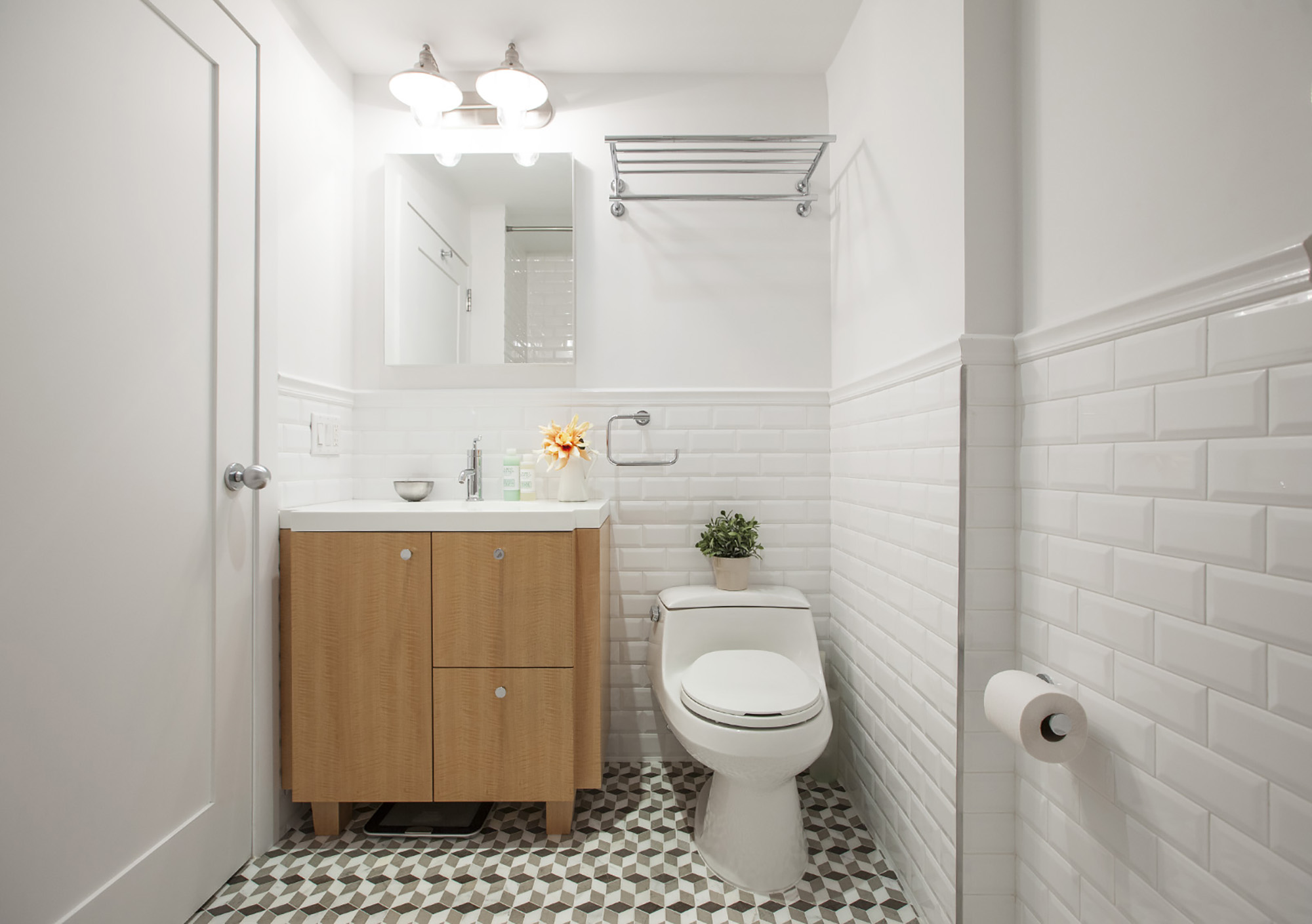 A renovated bathroom in Park Slope featuring white subway tile wall, geometric floor tile, wooden vanity, and overhead mirror light