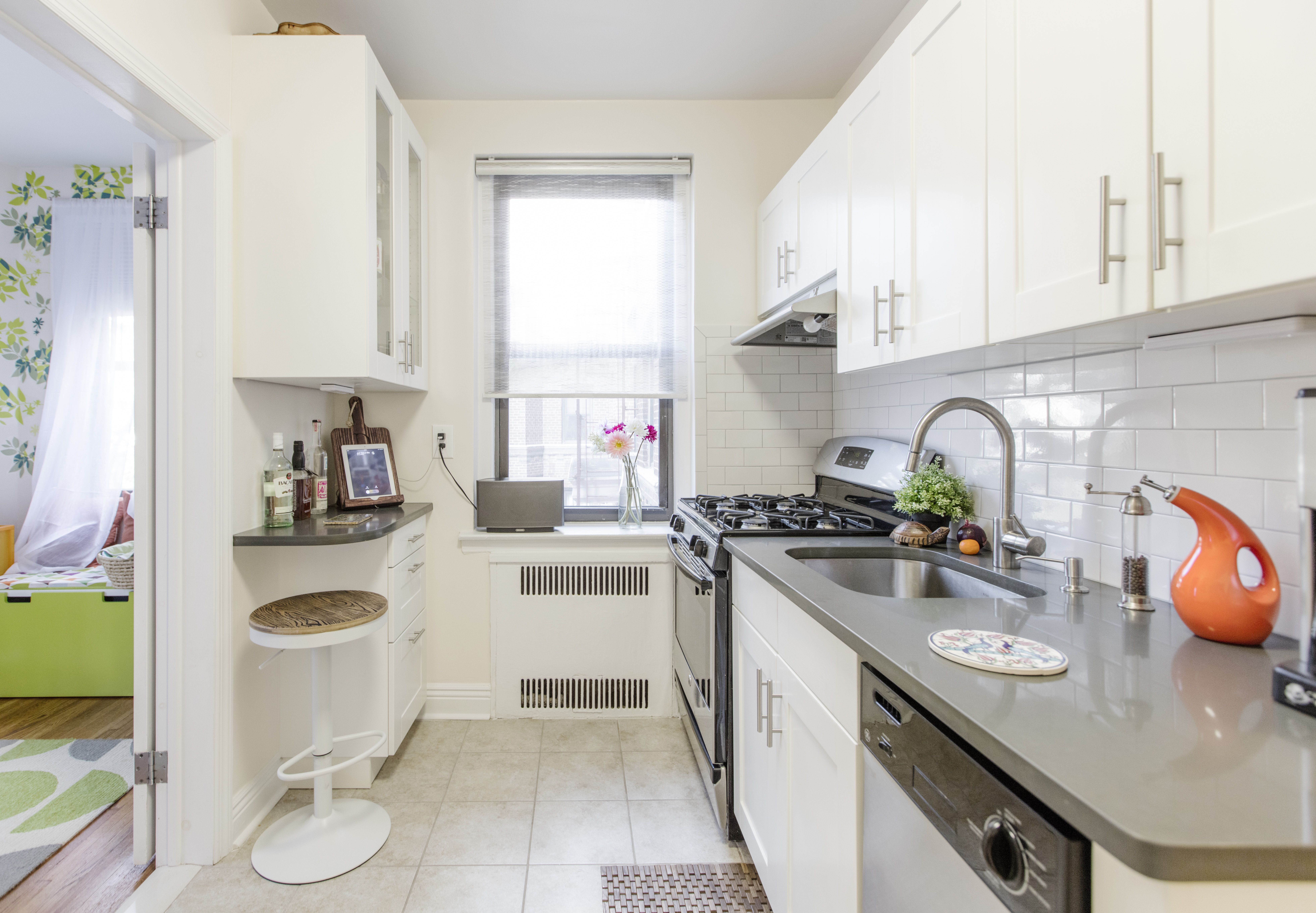 Remodelled Ditmas Park kitchen featuring white shaker cabinets, stainless steel appliances, and a built-in corner counter overlooking a bright adjoining room.