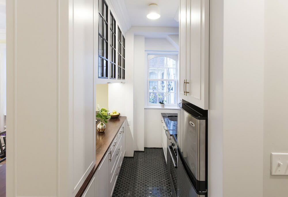 narrow kitchen with black floor tiles and flush mounted ceiling light and white walls and cabinets and butcher block countertop on pass through window after renovation