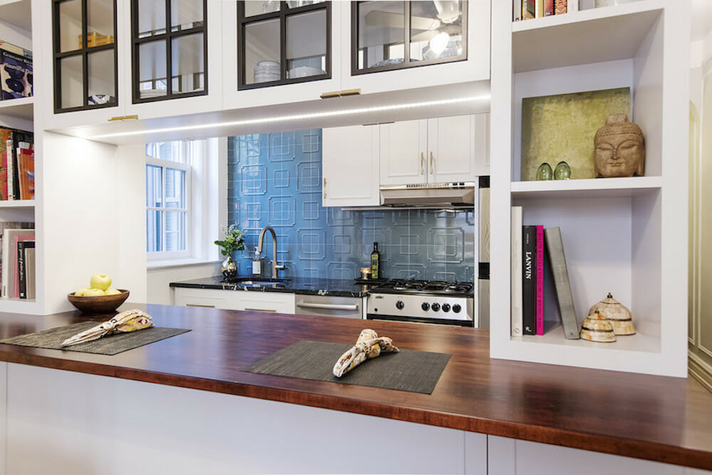 pass through window with butcher block countertop and overhead white cabinets with glass doors and storage on sides and kitchen with blue backsplash after renovation