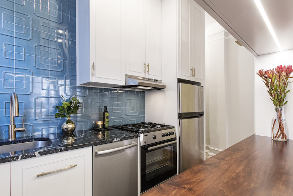 white kitchen cabinets with brass handles and black marble countertop with undermount sink and stainless steel appliances and blue backsplash and butcher block countertop on island after renovation
