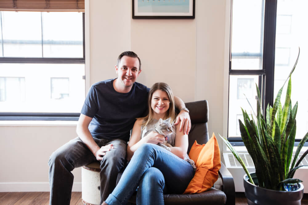 laura posing with her husband on a chair in living room with hardwood floors and windows with black frames after renovation