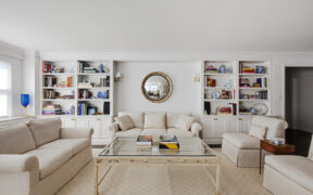 Upper East Side living room custom built-ins frame a renovated sofa nook with books, ceramics, brass sconces, pale seating, and a glass coffee table.