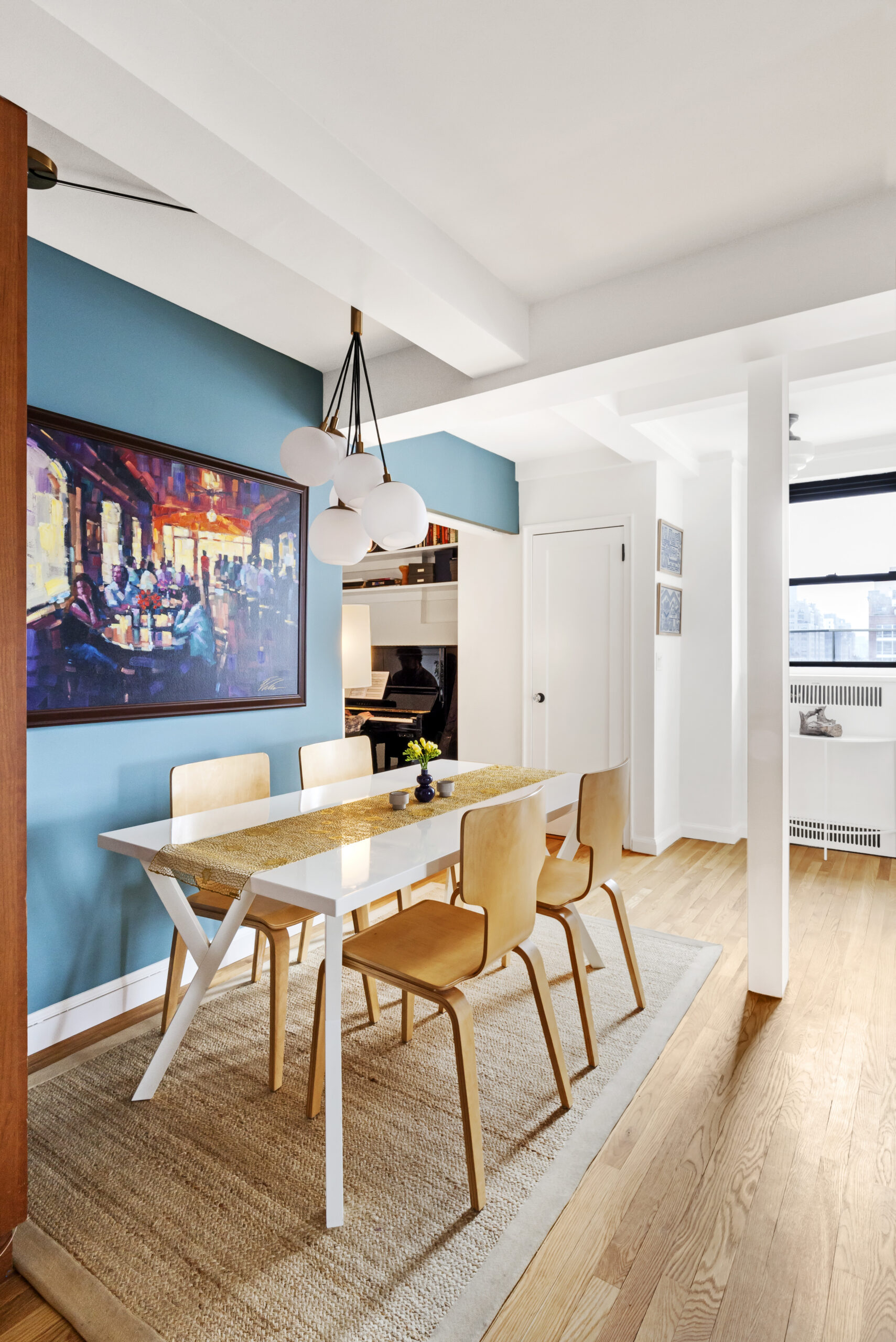 Chelsea dining area after renovation with Hamilton Blue wall, globe chandelier, refinished wood floors, artwork, and piano nook beyond.