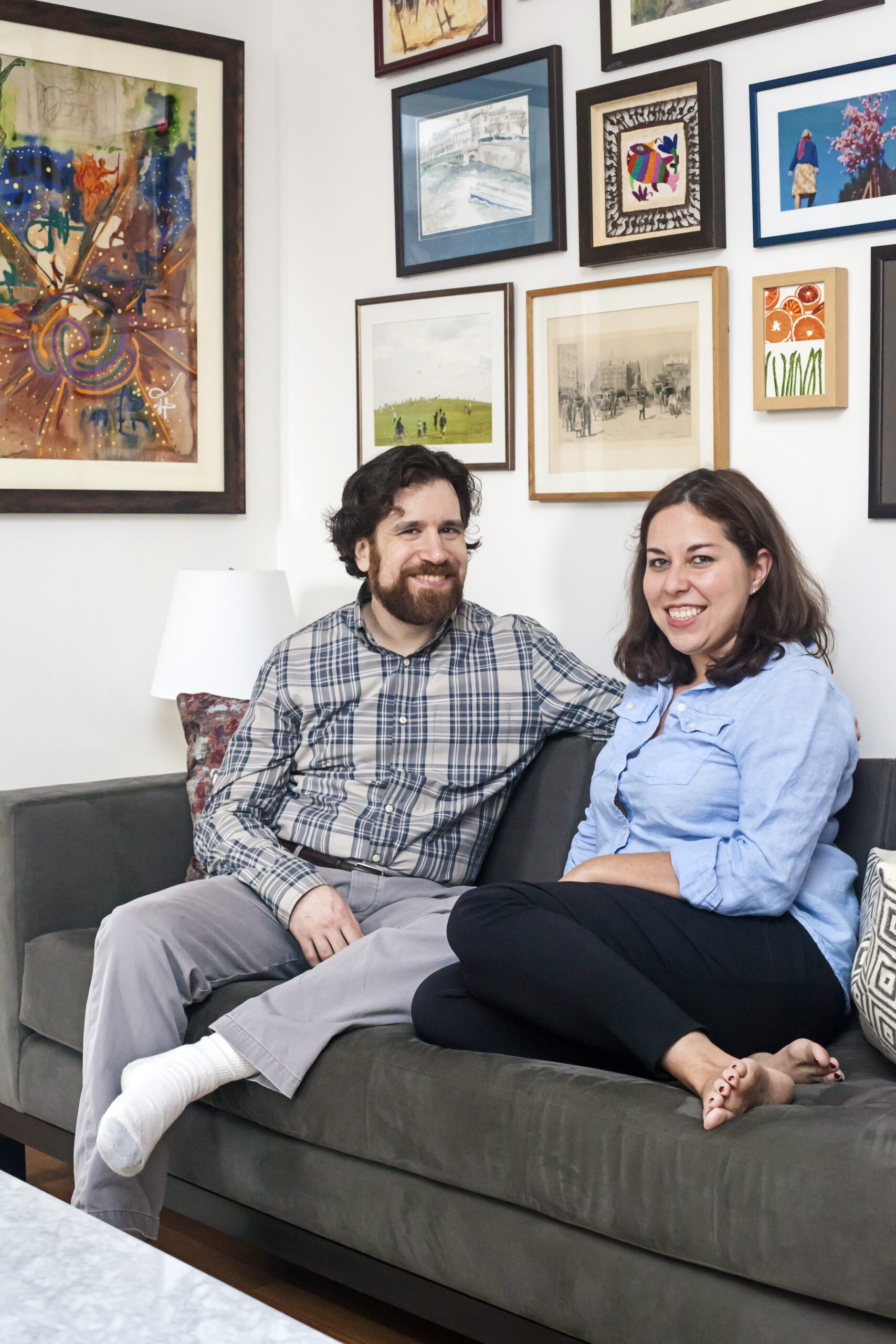 Chelsea homeowners seated on a gray sofa after renovation, with a colorful gallery wall, framed artwork, pillows, and a bright living area.