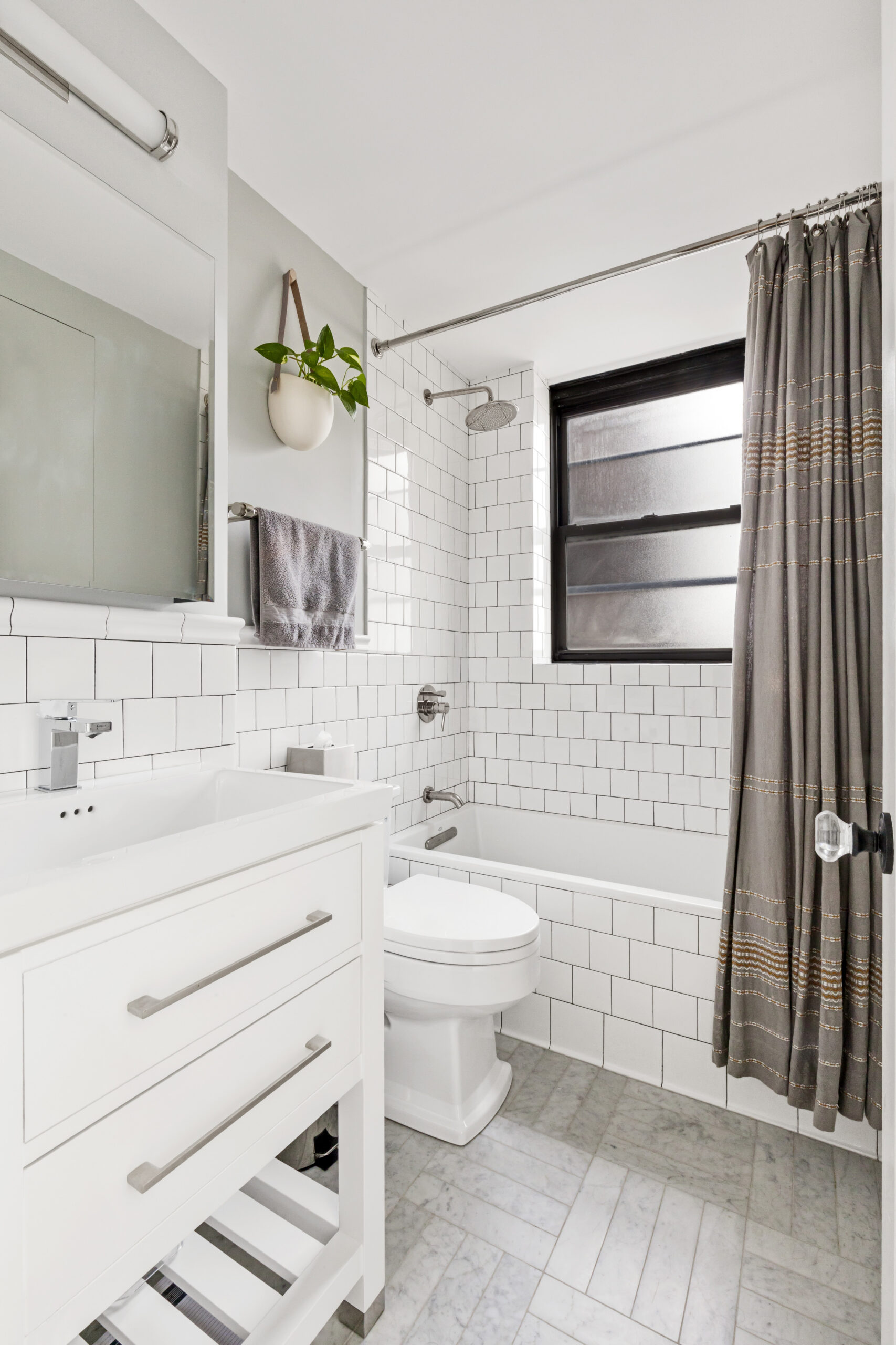 New York City guest bath with white subway wall tile, black grout, marble floor tile, white vanity, tub, shower curtain, and dark window trim.