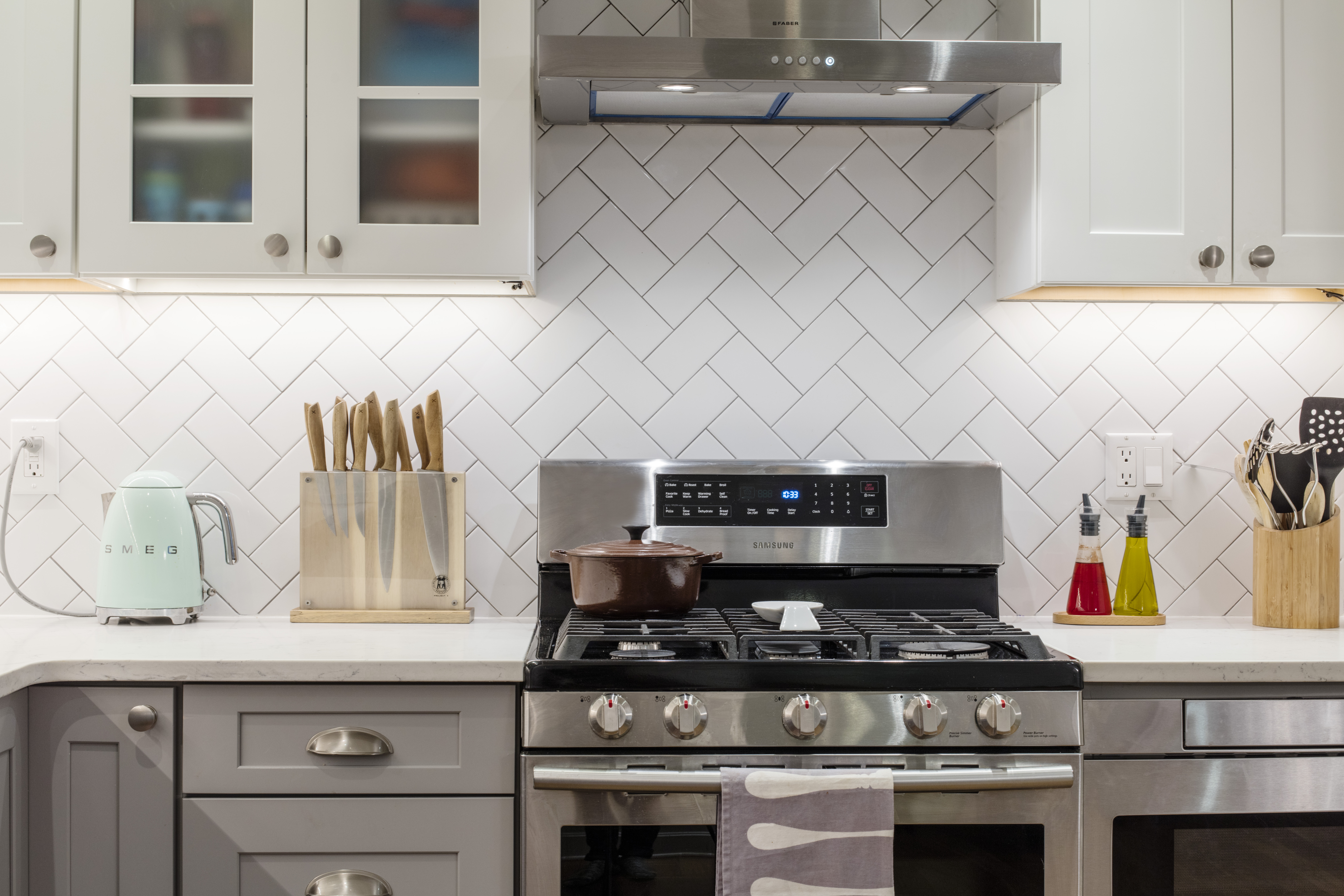 Brooklyn kitchen detail with white herringbone tile, stainless range hood, shaker cabinets, gray drawers, and marble-look counters.