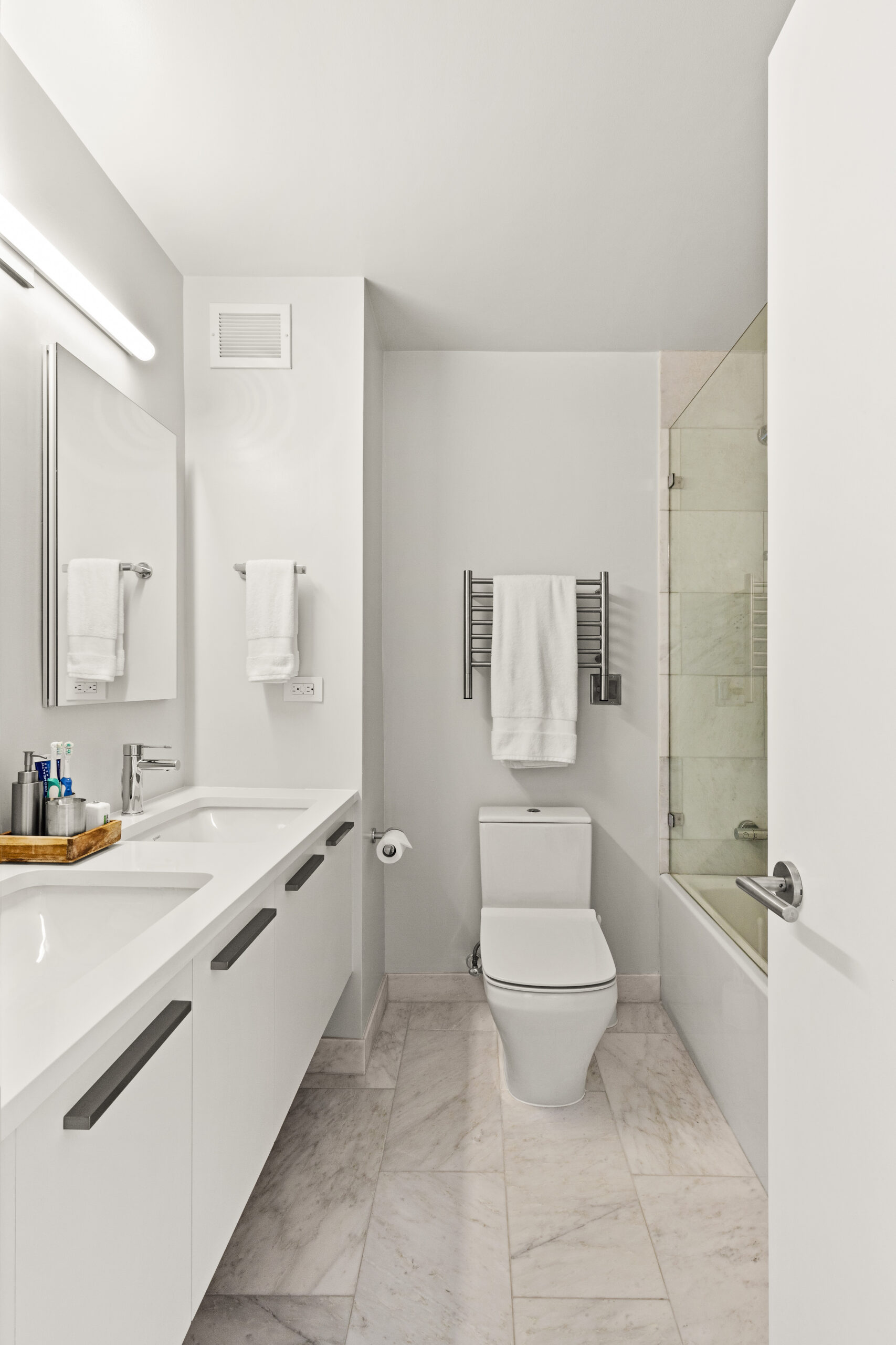 New York bathroom after renovation with a double vanity, two frameless mirrors, bright wall light, chrome faucets, and clean white finishes.