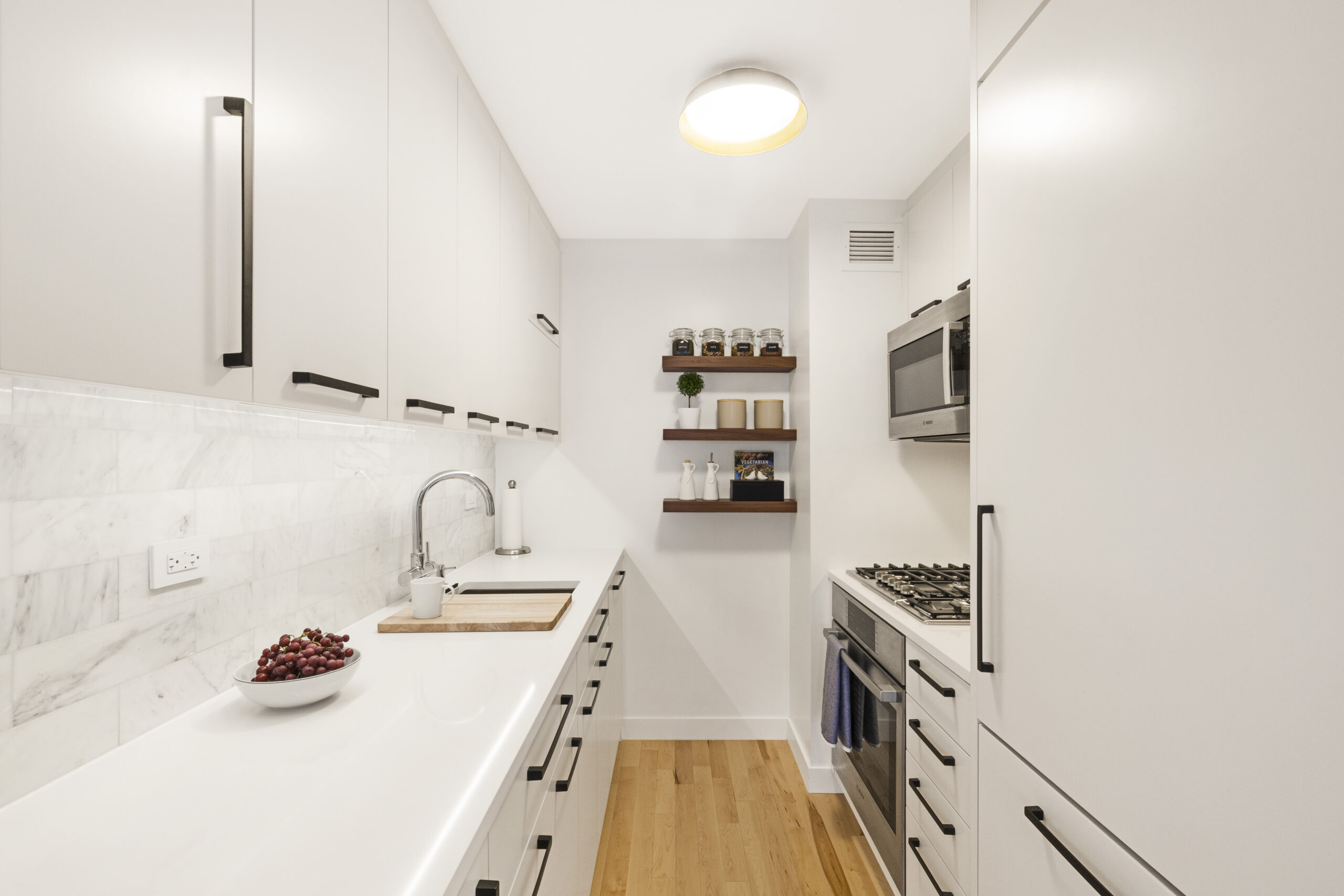 New York galley kitchen after renovation with white cabinets, marble subway tile, quartz counters, black pulls, wood floors, and open shelves