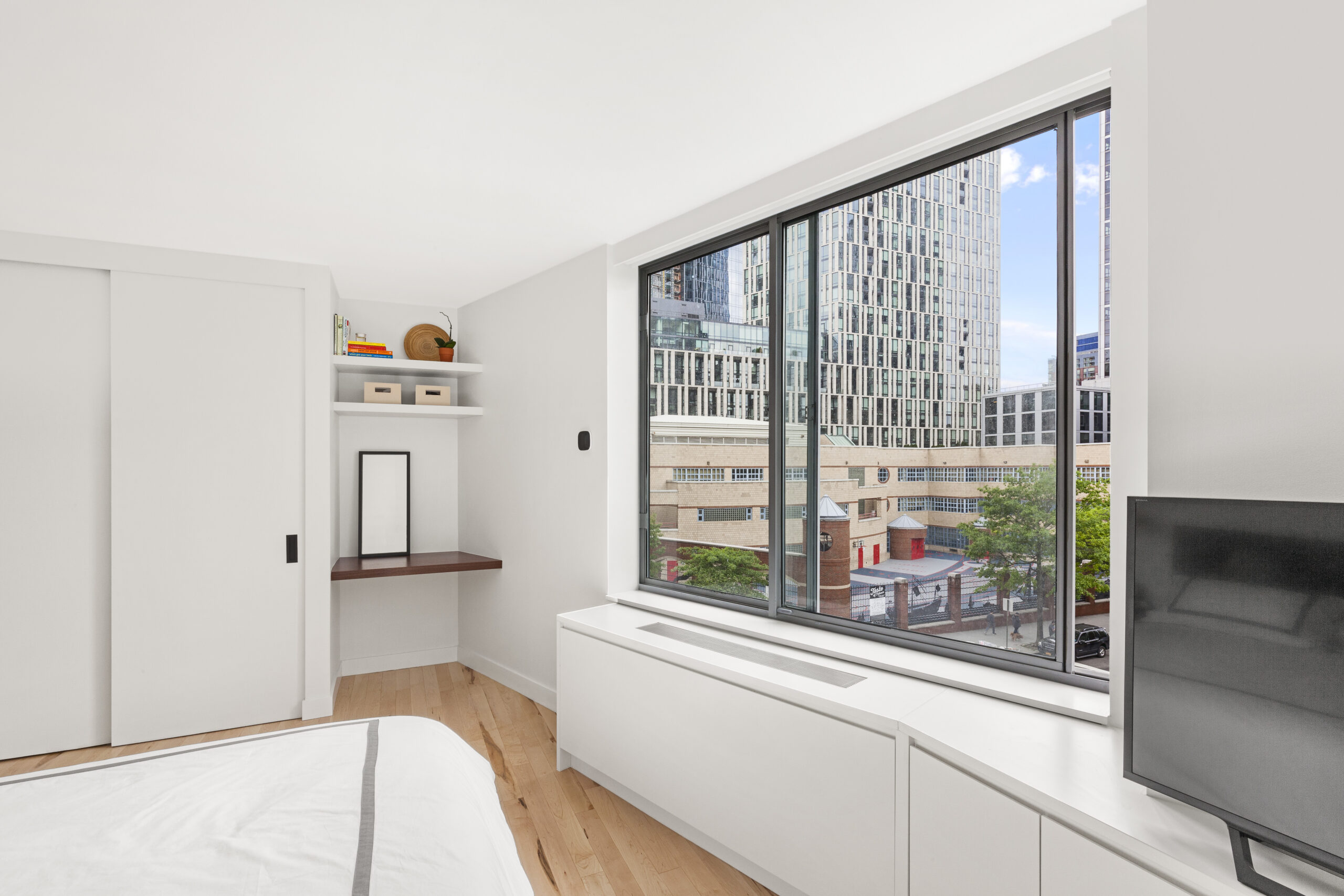 New York bedroom after renovation with sliding closet doors, white built-ins, a wood vanity, large windows, light wood floors, and a TV wall.