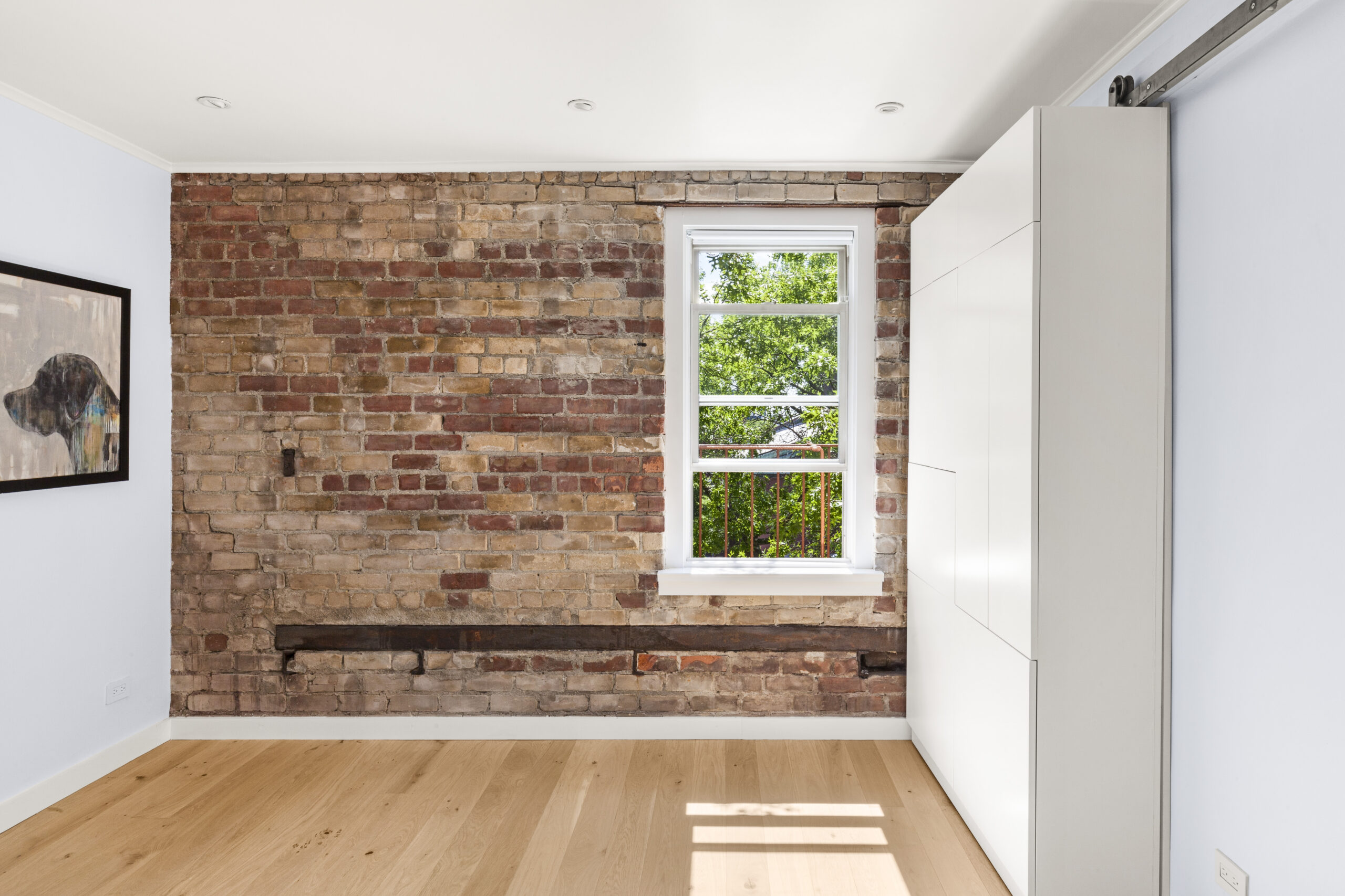 Cobble Hill co-op bedroom featuring an exposed brick wall, original horizontal beam, white trim, pale wood floors, and a sliding custom wardrobe.