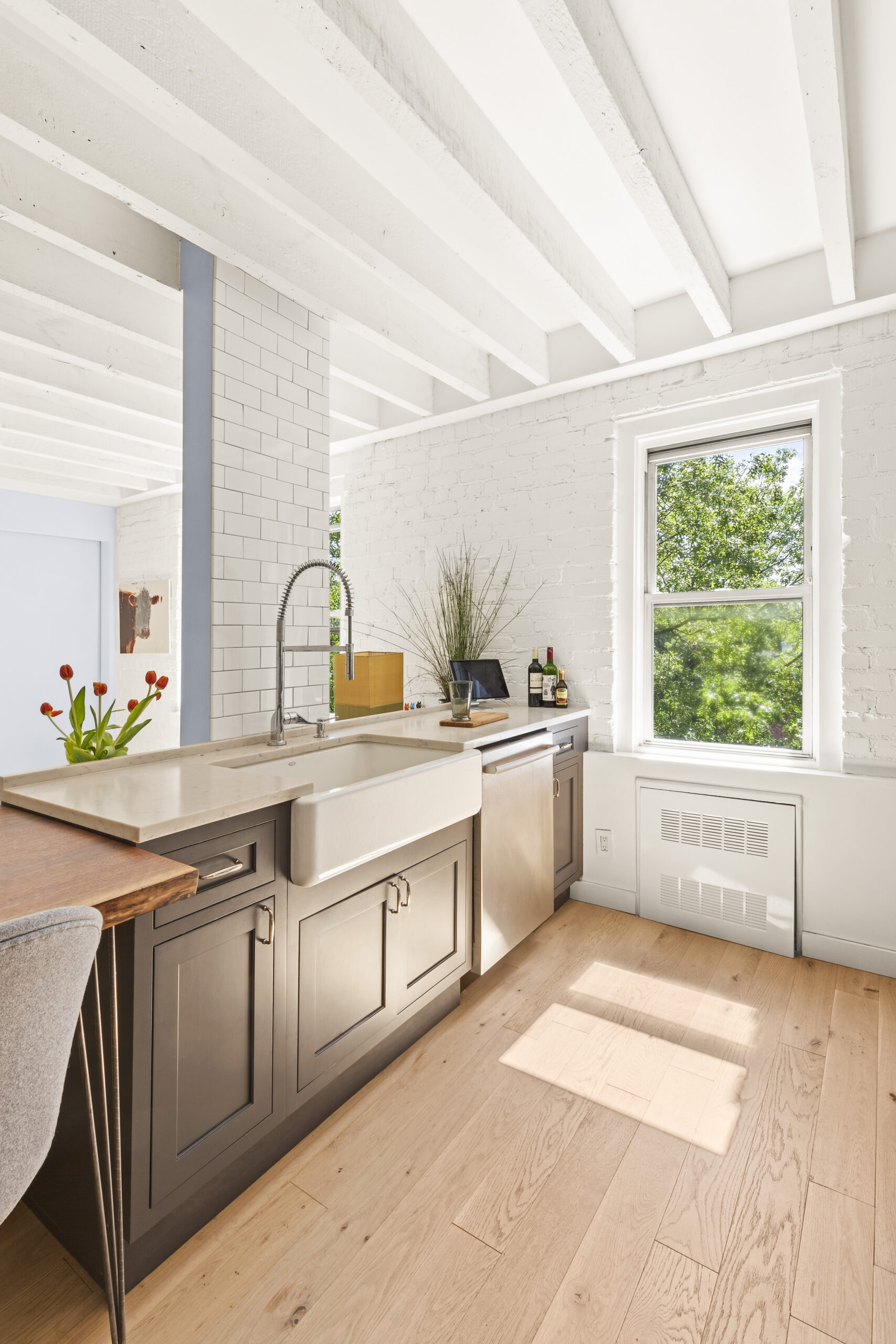 Cobble Hill co-op kitchen with gray cabinetry, a white farmhouse sink, quartz counters, subway tile, exposed beams, and engineered wood flooring.