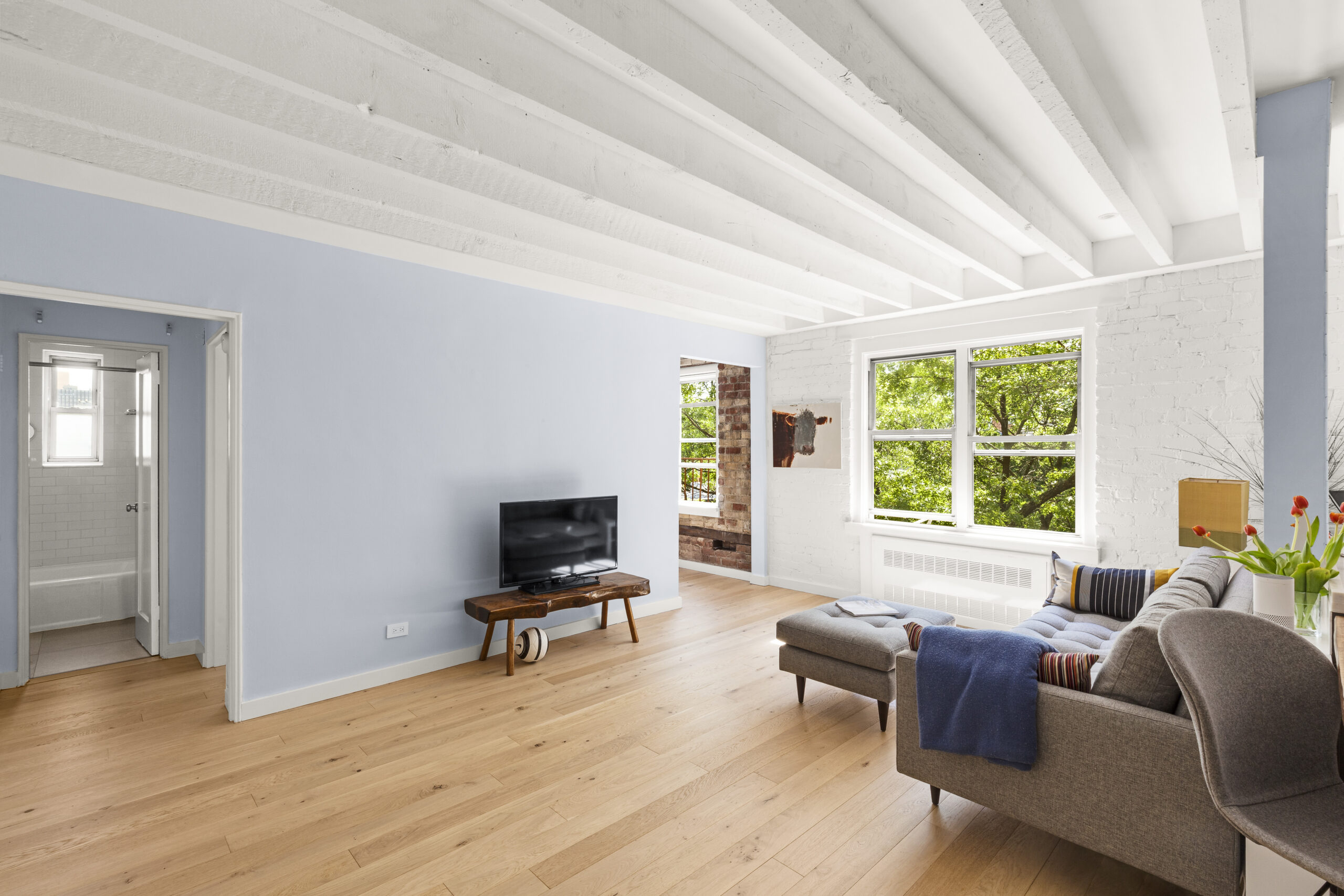 Cobble Hill co-op living room with engineered wood floors, painted brick, white ceiling beams, blue-gray walls, and widened openings to adjoining rooms.