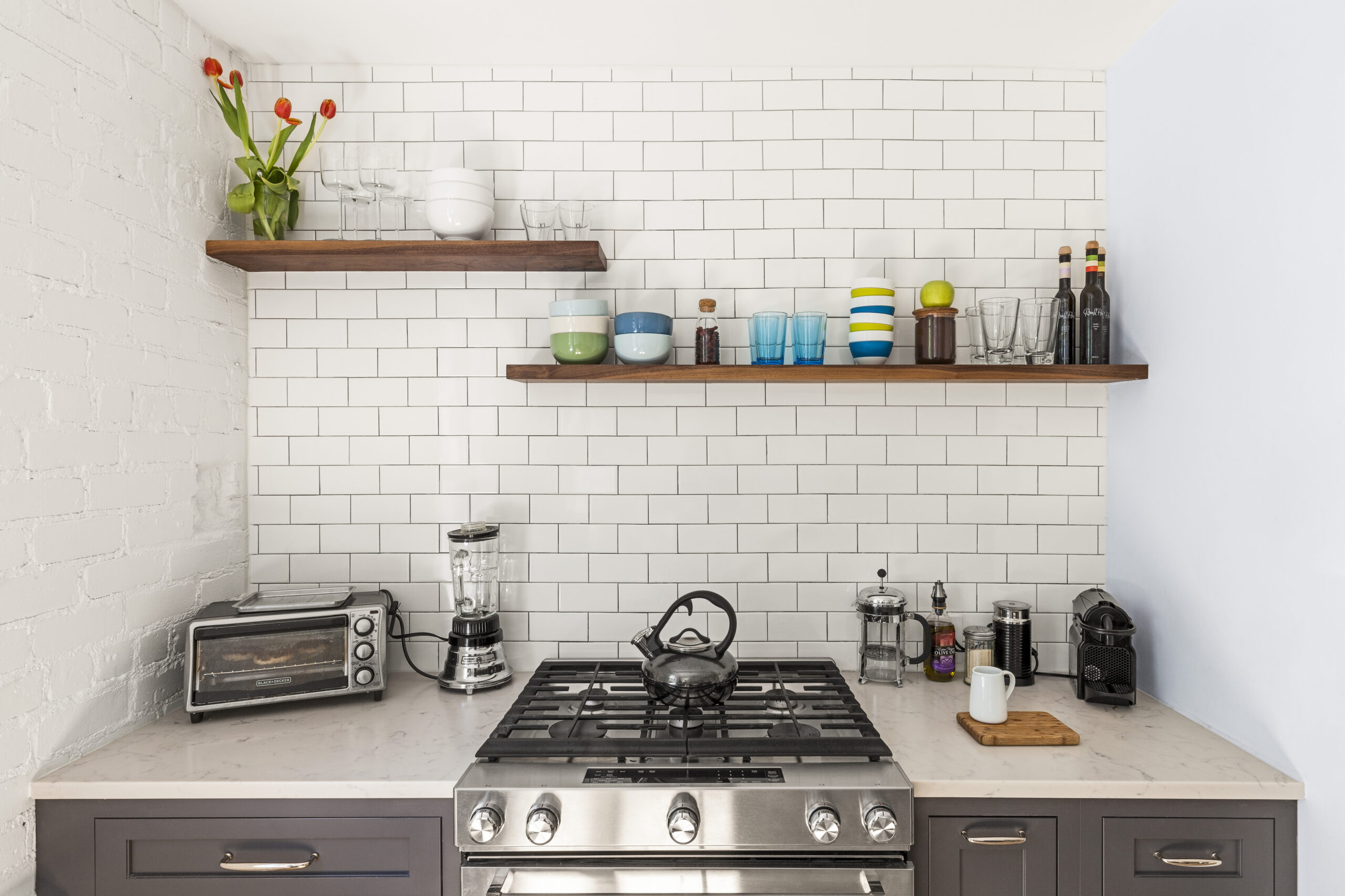 Cobble Hill co-op kitchen with charcoal base cabinets, stainless range, quartz counters, white subway tile, and walnut open shelves for everyday storage.