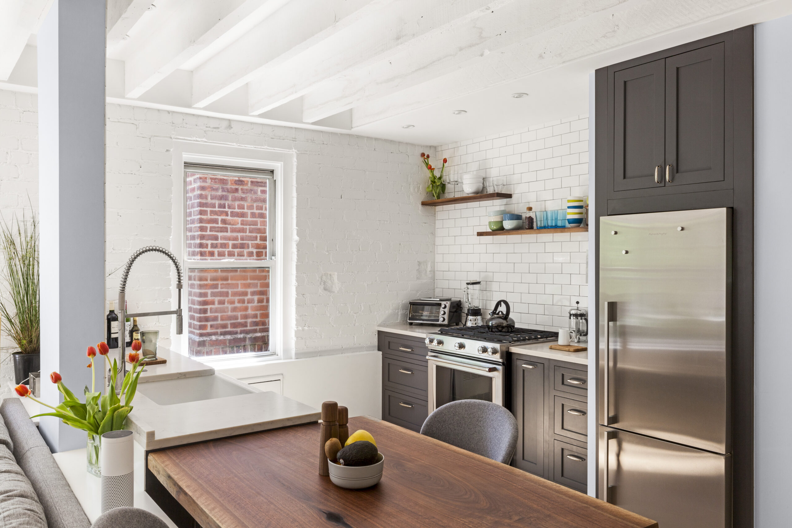 New York City co-op kitchen with charcoal cabinets, white subway tile, open walnut shelves, stainless appliances, quartz counters, and a movable dining table.