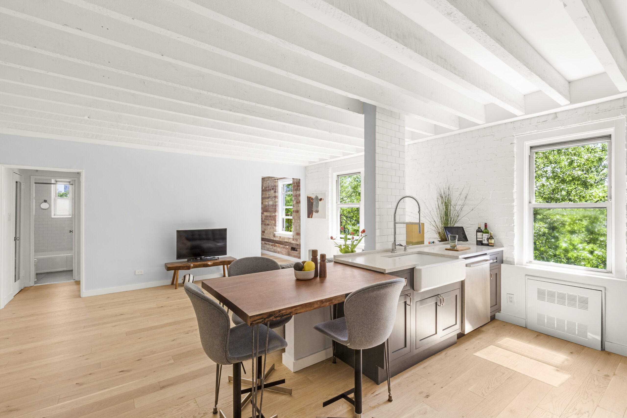 New York City co-op renovation with a movable walnut dining table, farmhouse sink, gray cabinets, exposed beams, painted brick, and open living space.