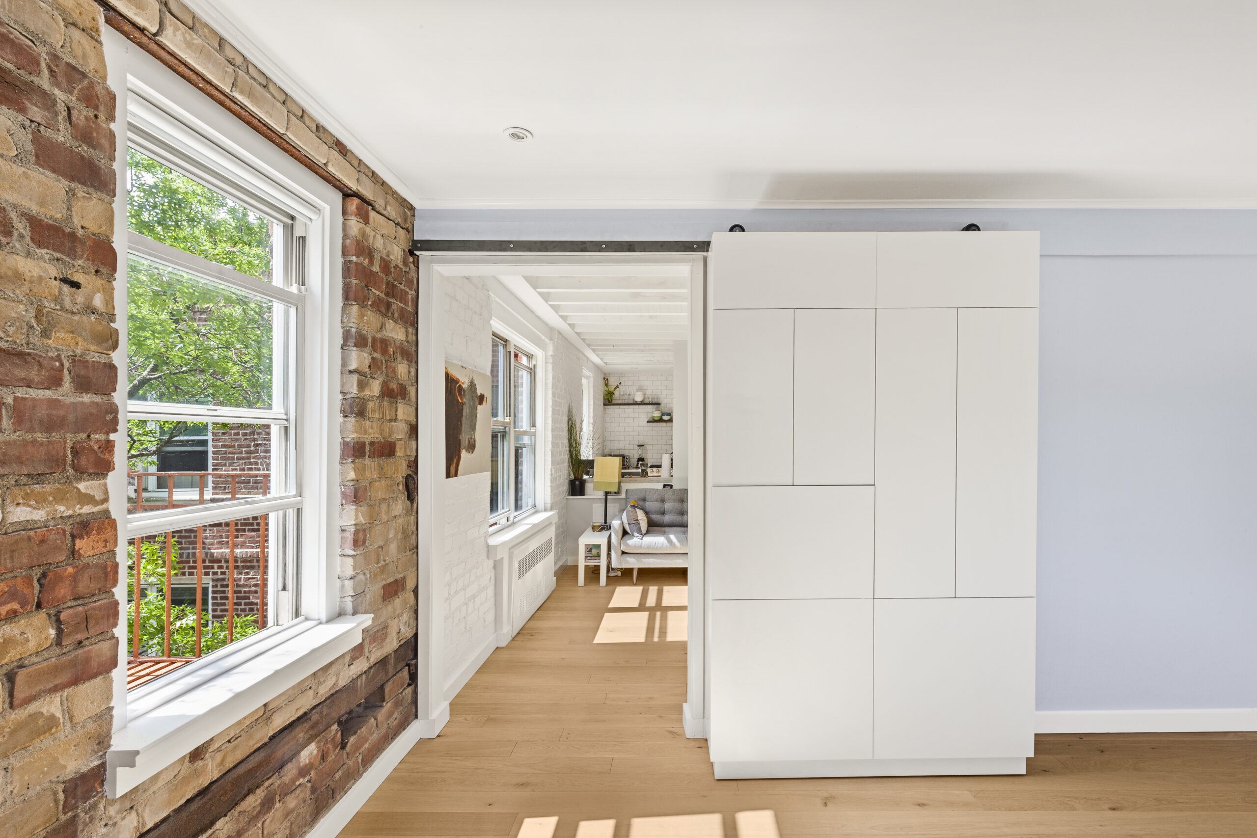New York City co-op bedroom with exposed brick, pale wood flooring, and a custom white wardrobe mounted on a sliding track beside the widened doorway.