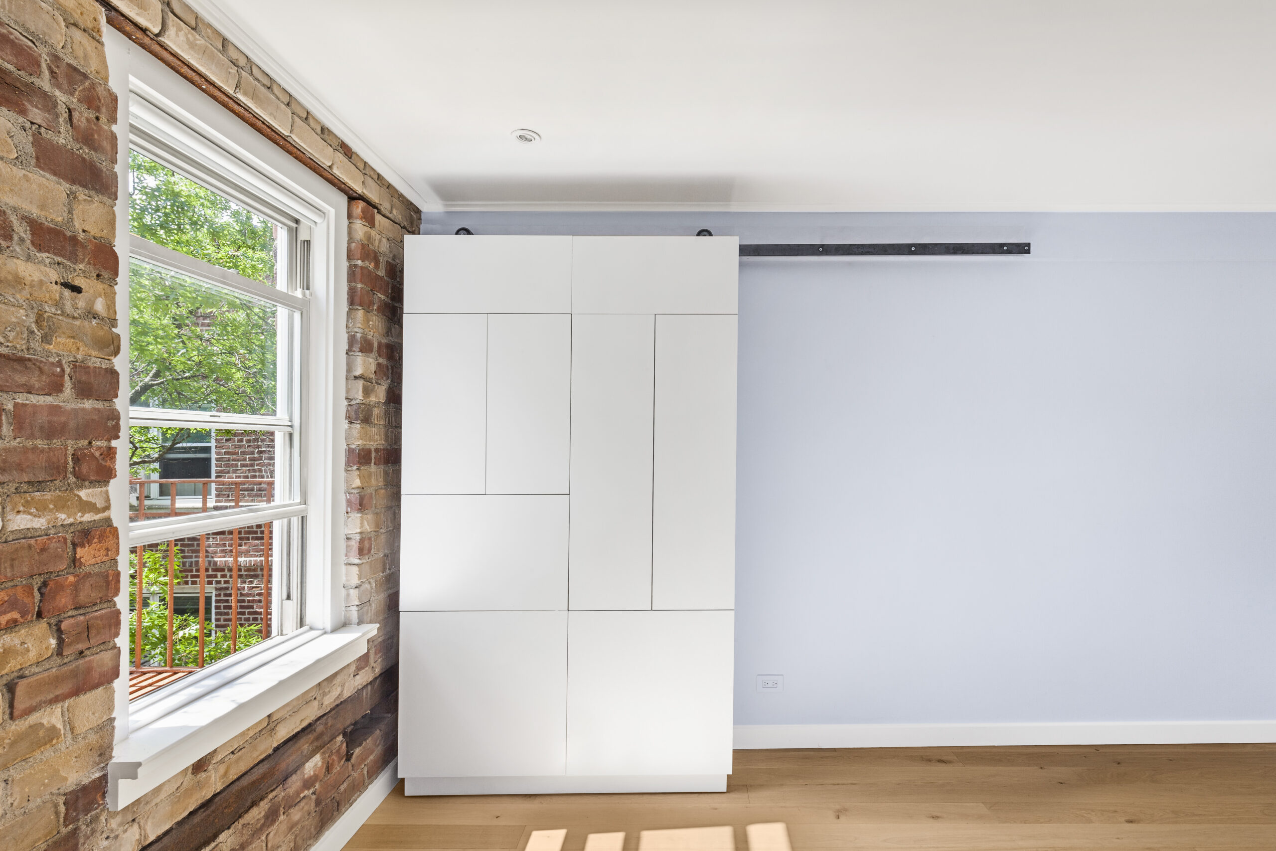 New York City co-op bedroom with a floor-to-ceiling white wardrobe on a black metal track, exposed brick by the window, and engineered wood floors.