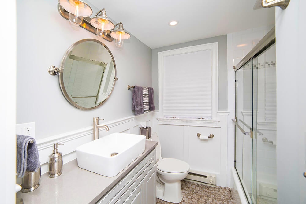 White and gray bathroom with farmhouse sink plus vanity under a round vanity mirror and light fixtures after renovation