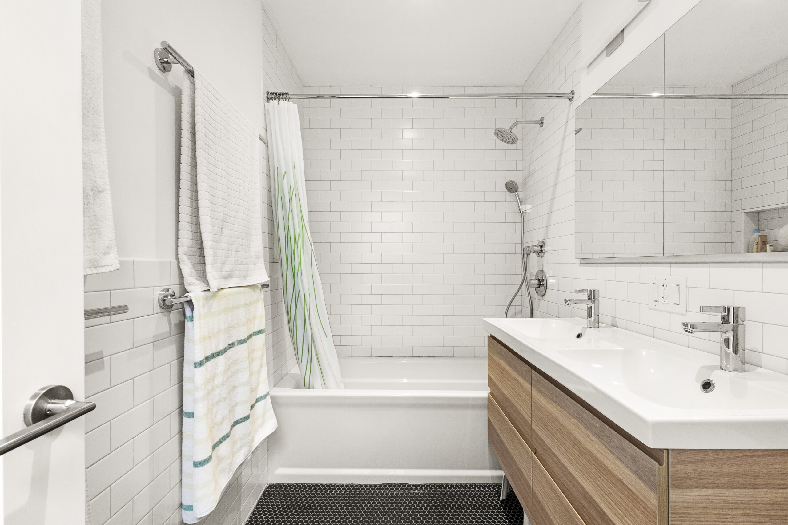 Bedford-Stuyvesant bathroom with white subway tile, black hex floor tile, a wood double vanity, mirrored cabinet, and a tub-shower combo.