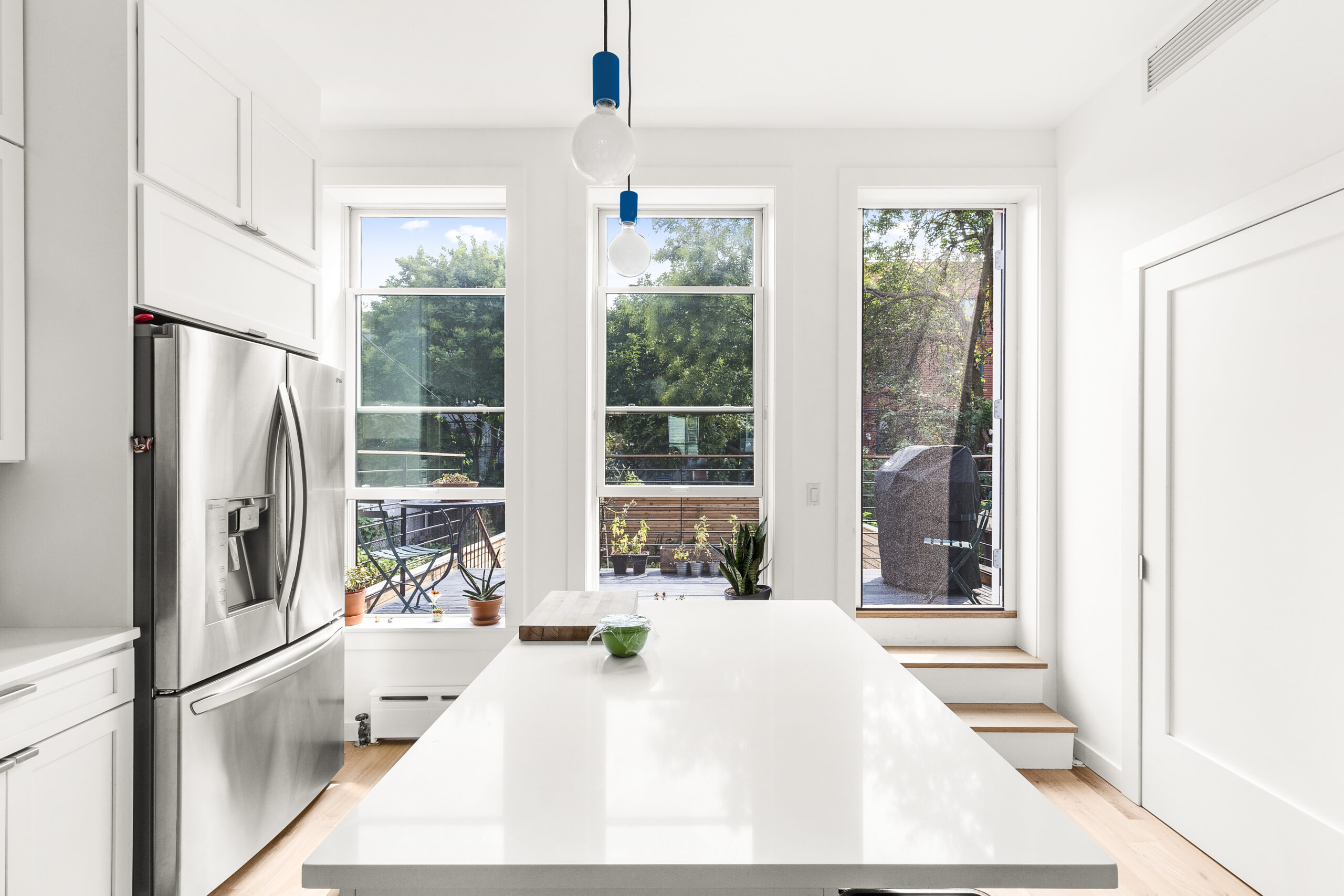 White Oak Wood Flooring brightens this Brooklyn kitchen, with a white island, rear windows, pendant lights, and steps leading to the deck.