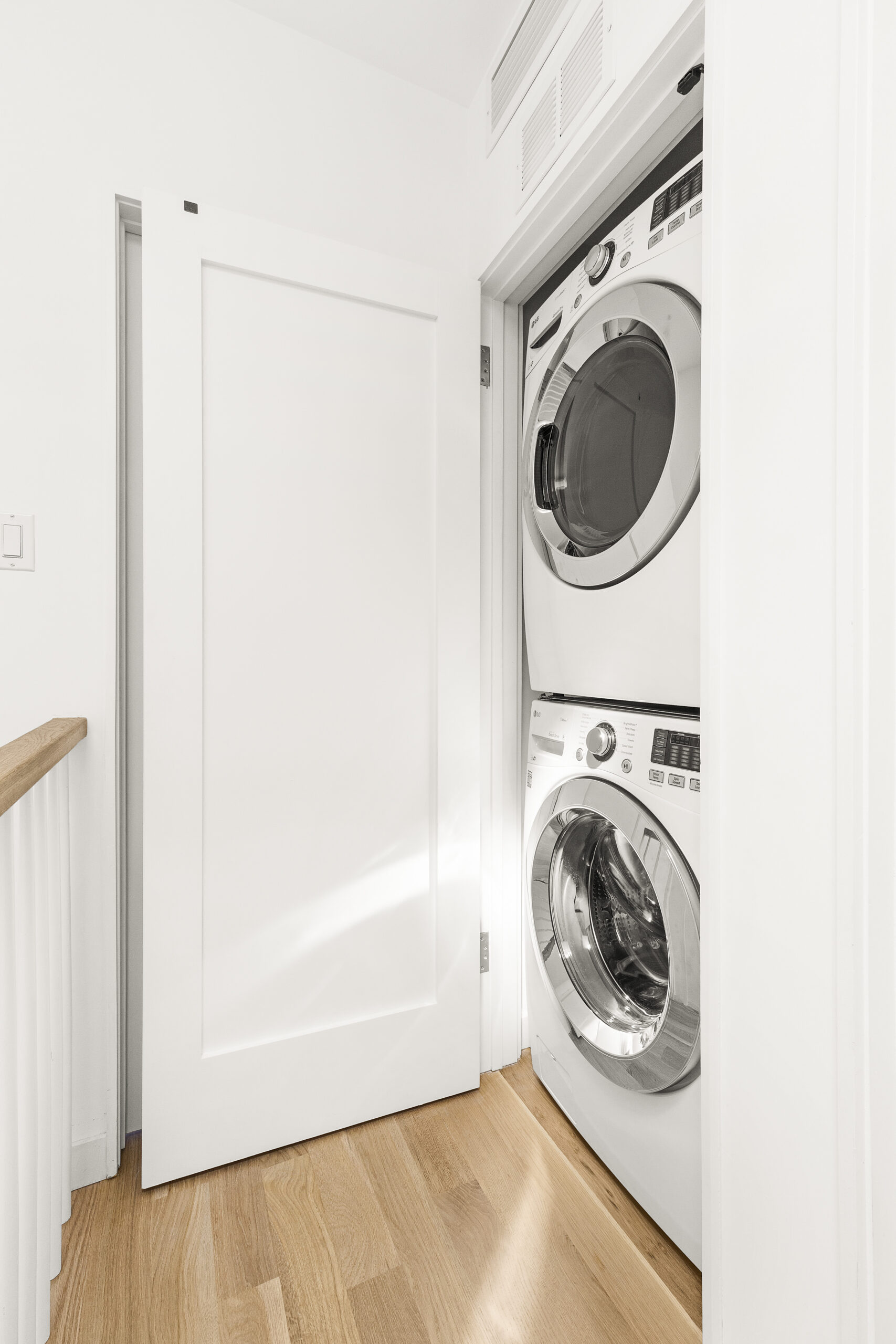 White Oak Wood Flooring leads to a Brooklyn laundry closet with a stacked washer and dryer, white walls, and a clean door tucked off the hall.