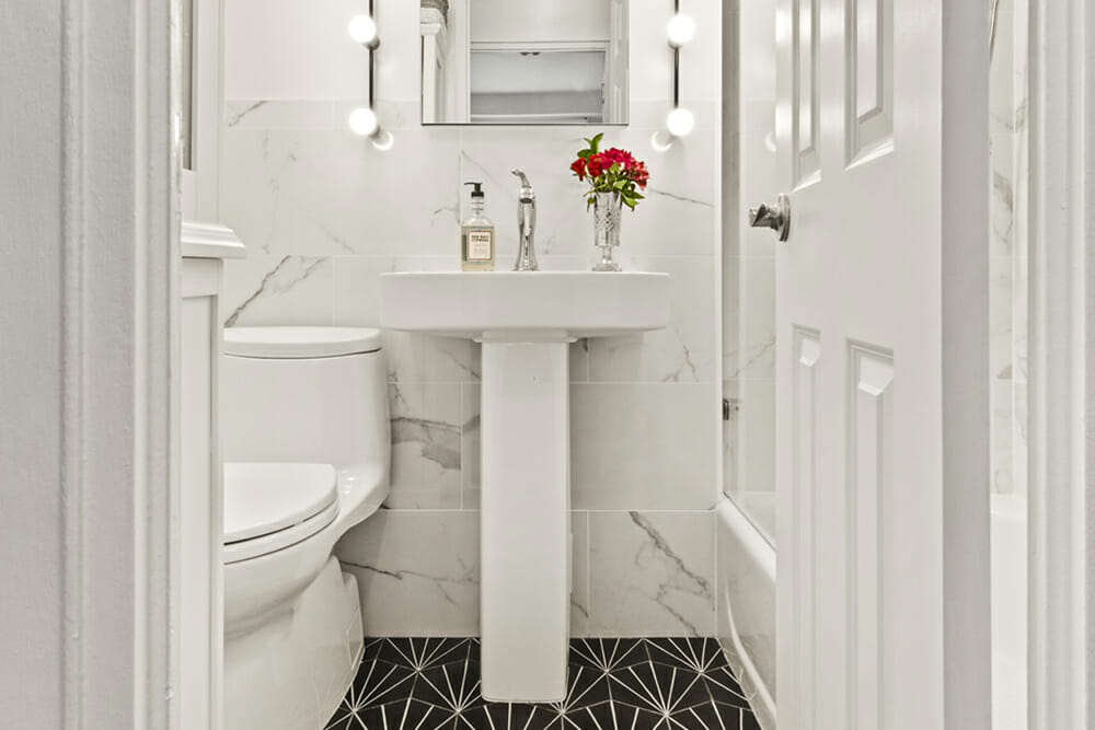 white pedestal sink in a white bathroom with white marble wall and white door with black patterned floor after renovation