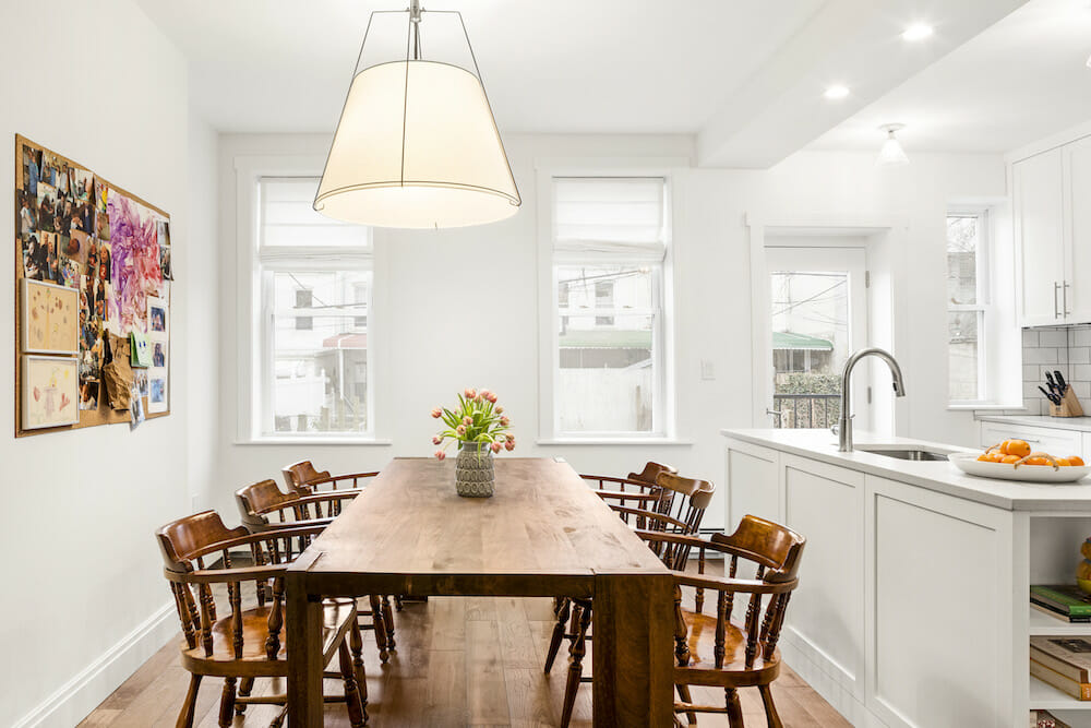 natural brown dining table with chairs in a full white kitchen with white kitchen cabinets and two long windows after renovation 
