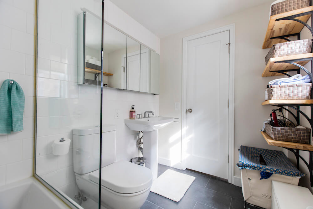 white bathroom with gray floor tiles and bathtub with glass wall and floating wooden storage shelves with iron brackets after renovation