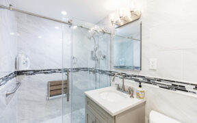 Walk-in shower with glass doors, grab bars, and a fold-down seat beside a vanity, showing senior-friendly bathroom design in white and gray.