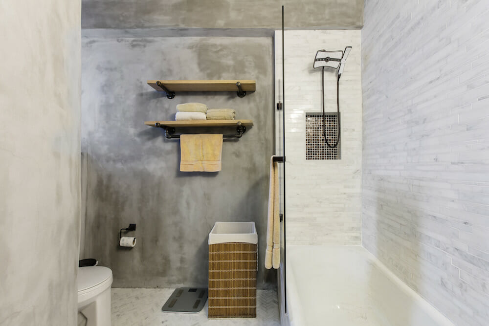 concrete bathroom walls with floating wooden shelves and toilet and floor tiles and bathtub with glass wall and light gray tiles on wall after renovation