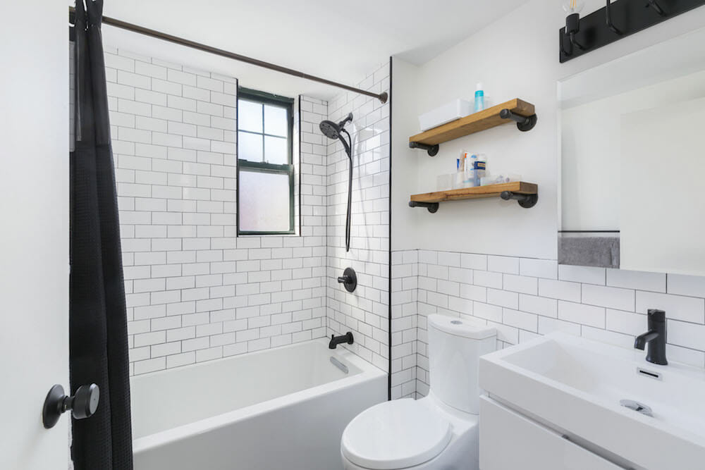 white subway tiles with black grout and black shower head and fixtures and bathtub and floating wooden shelves above toilet and vanity with mirror after renovation