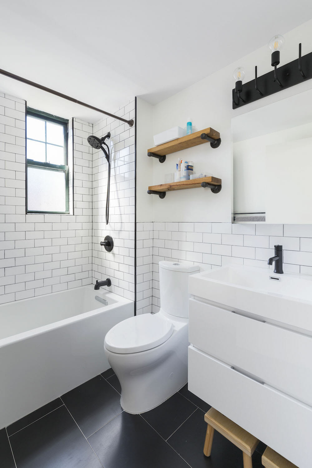 white subway tiles with black grout and black shower head and fixtures and bathtub and floating wooden shelves above toilet and floating vanity and black floor tiles after renovation