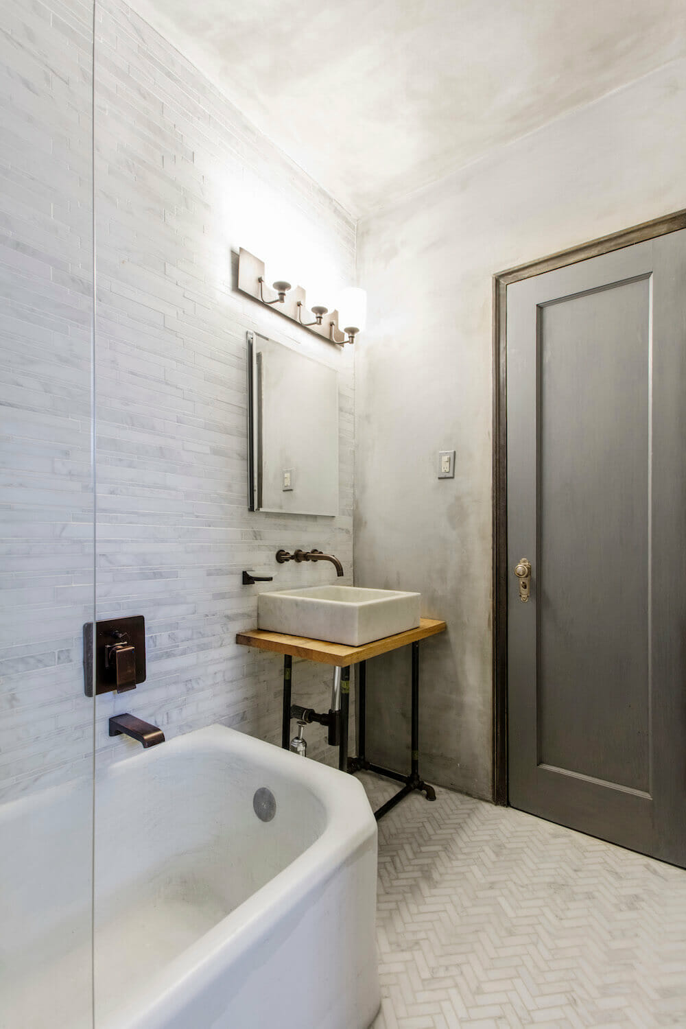 Gray door in a white bathroom with white bathtub and herringbone floor tile after renovation