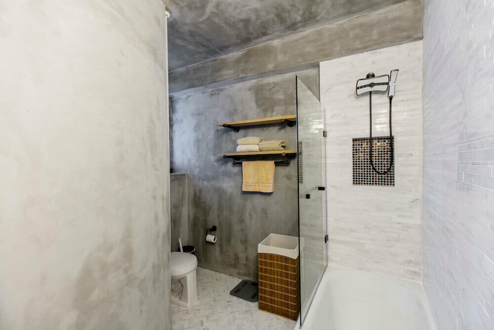 Gray cement wall and white bathtub with tempered glass separator and floating wall shelf after renovation