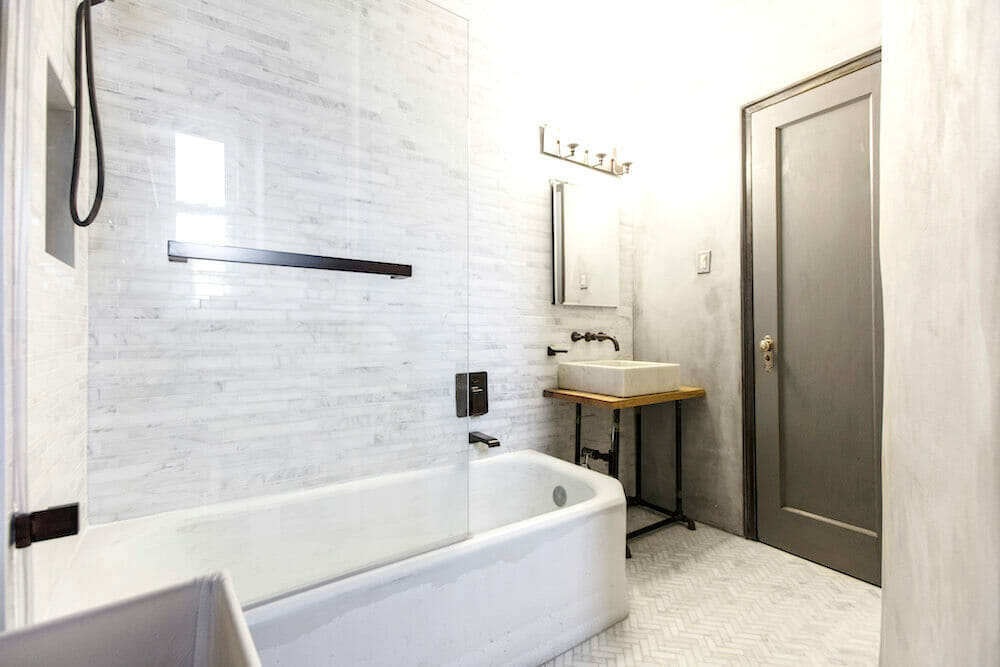 Long white tub with white wall tile and marble herring bone floor tile after renovation