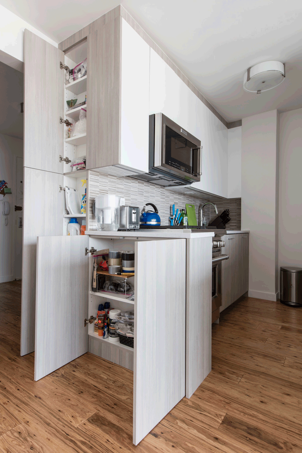 white overhead kitchen cabinets and light gray under counter cabinets with white countertop and gray backsplash tiles and hardwood floors and flush mounted light after renovation