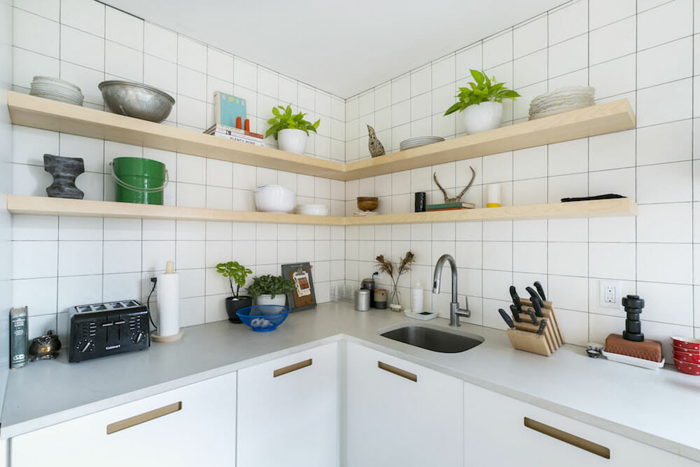 white kitchen cabinets with flush pull handles and gray countertop with undermount sink and white square backsplash tiles and wrap around floating wooden corner shelves after renovation