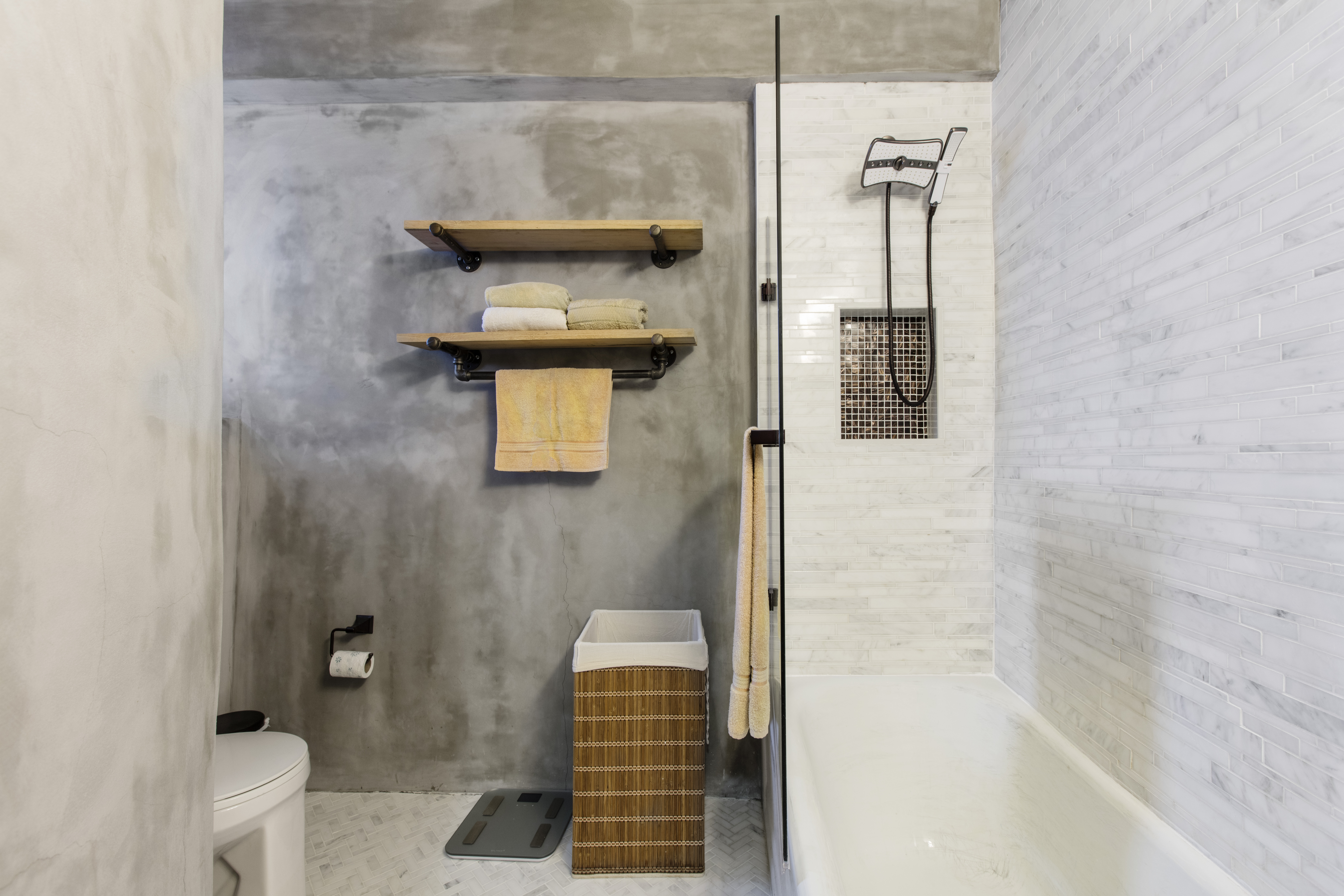 Wabi-sabi bathroom in Windsor Terrace with raw cement walls, reclaimed wood shelves, marble tile, a glass tub screen, and a contrasting shower niche.