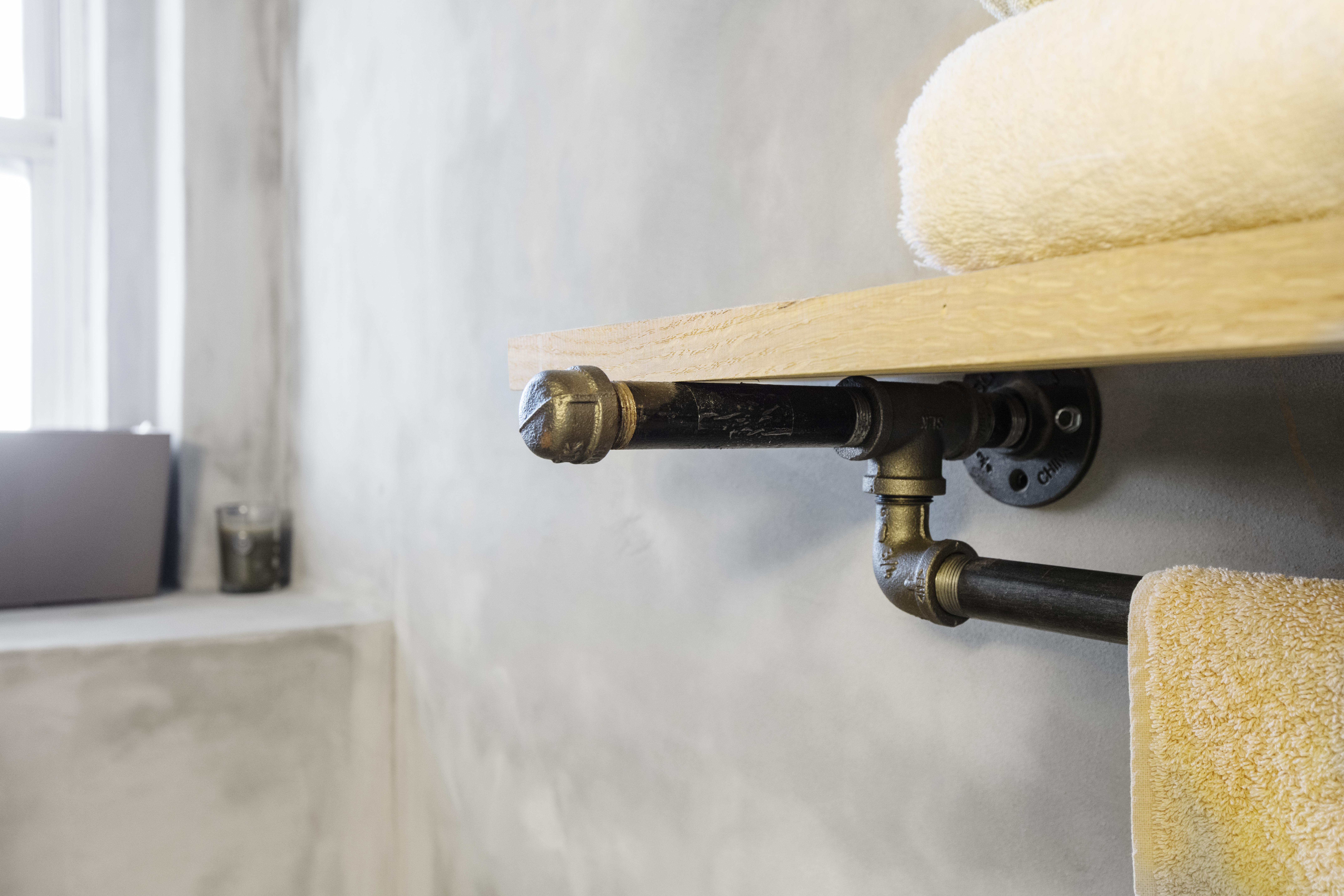 Brooklyn wabi-sabi bathroom detail with a reclaimed wood shelf, exposed pipe brackets, a built-in towel rail, soft linens, and textured cement walls.
