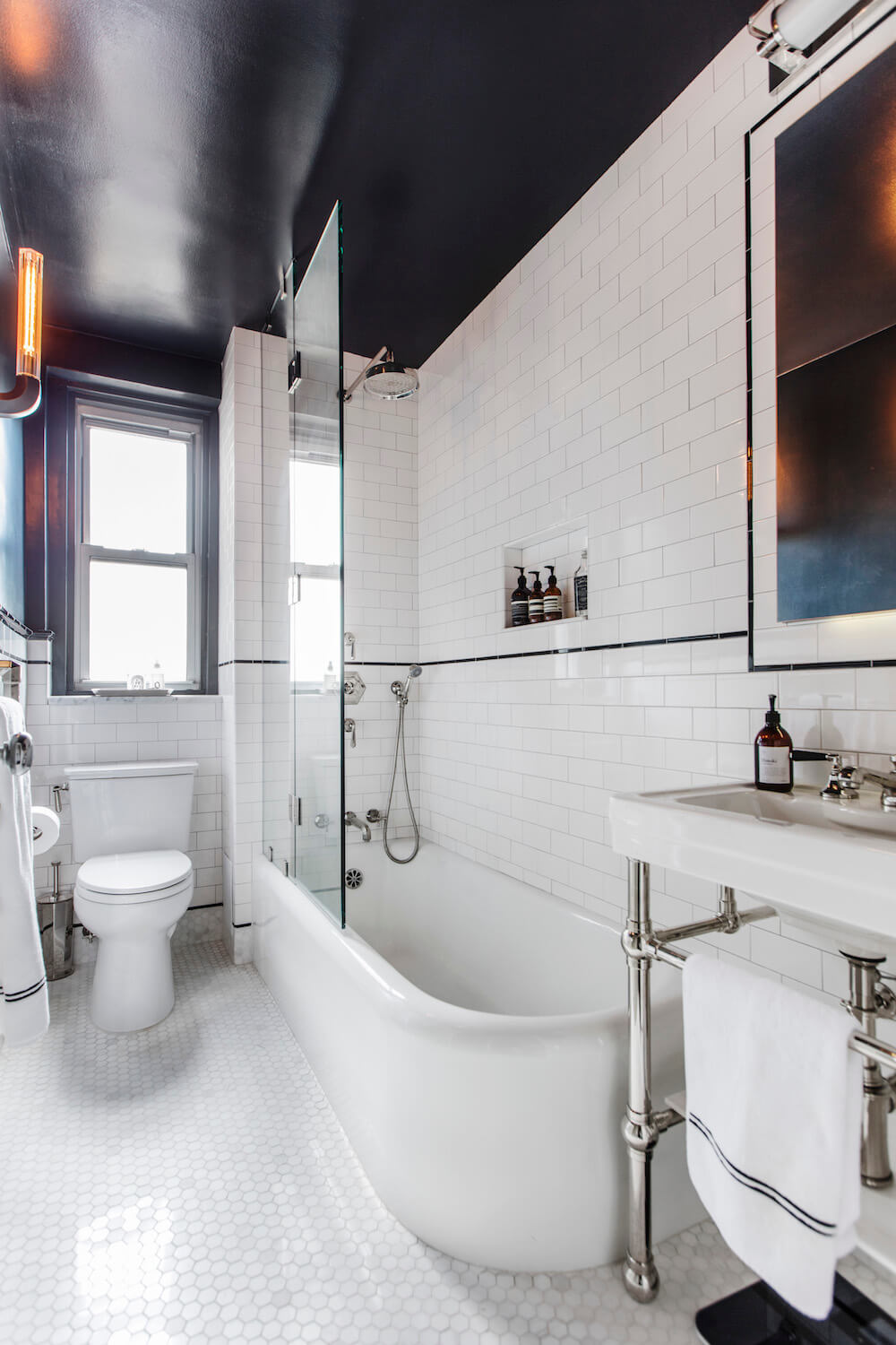 Black ceiling with white subway tiles in bathroom with white tub and shower glass and white hex floor tiles after renovation