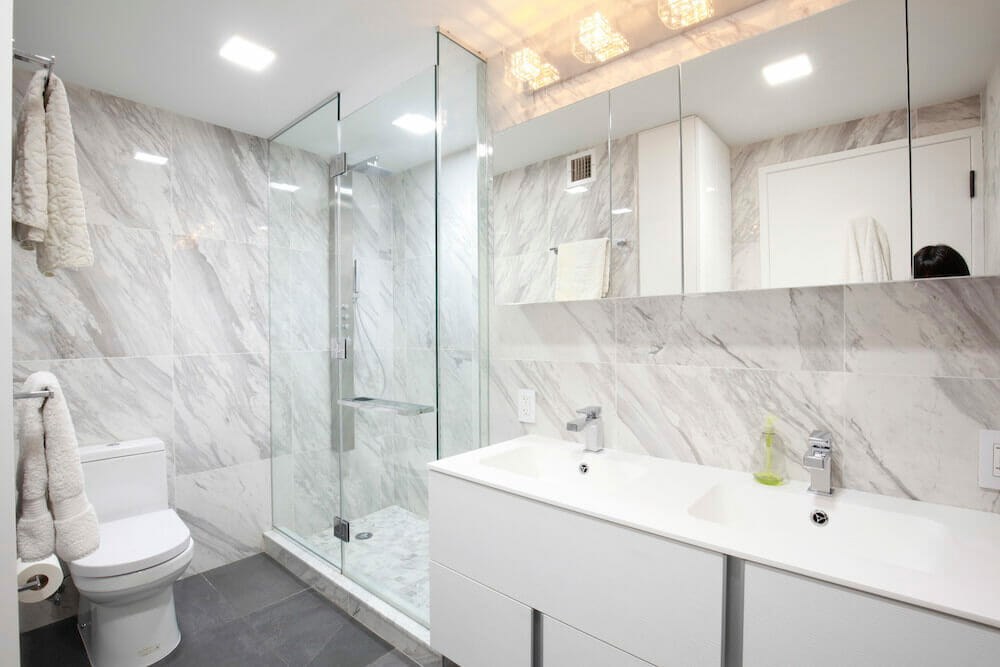 marble walls in a white bathroom with frameless glass doors and white double sink after renovation